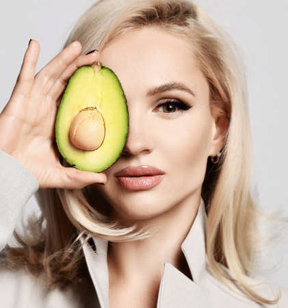 Pretty woman with blonde wavy hair and healthy skin wearing beige shirt holds half an avocado covering half of faceの写真素材