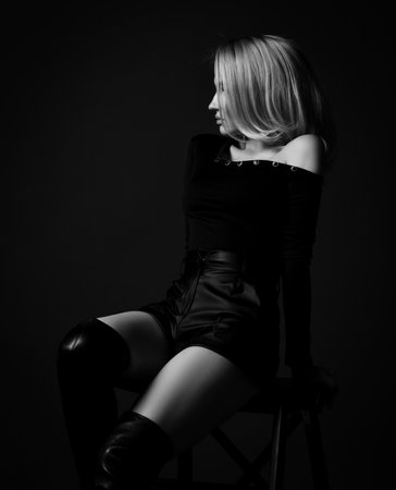 Profile silhouette of young slim pretty blonde woman in black sexy clothes and high boots sitting on stool looking asideの写真素材