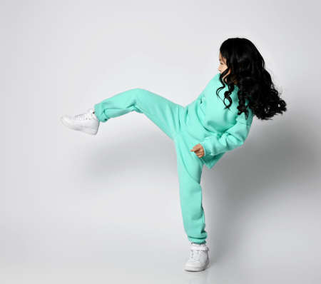 Frolic asian kid girl in pastel green, mint color hoodie and pants stands sideways kicking, playing footballの写真素材