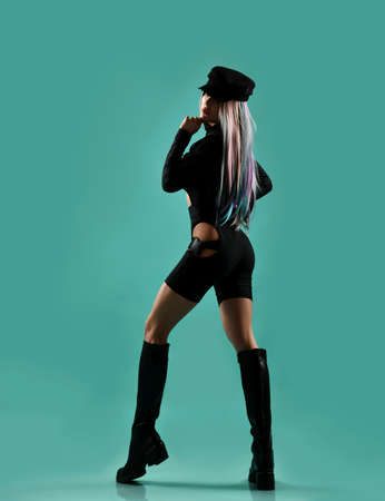 Young blonde woman with colored strands in black leather sexy costume and high boots standing...