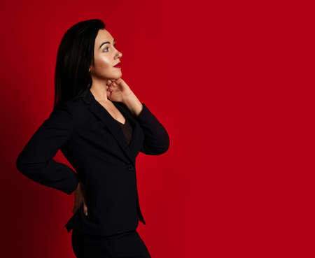 Slim adult woman in black official pantsuit stands sideways holding hand at her neck and looking at copy spaceの写真素材