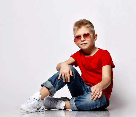 Cool blonde kid boy leader in red t-shirt, blue jeans, white sneakers and sunglasses sits on floor...