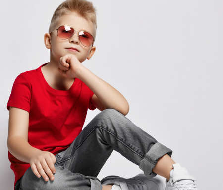 Cheerful stylish boy in red t-shirt, jeans, sneakers and sunglasses sitting on floor holding chin...