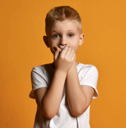 Young school boy kid in white t-shirt stands scared, eyes wide opened in surprise, covering his mouth with both handsの写真素材