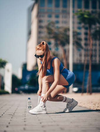 Athlete slim blonde woman trainer runner in blue sportswear ties her sneakers before workout on city...