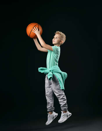Blond boy in green t-shirt, pants, sneakers and hoodie around waist jumps throwing the basketball ballの写真素材