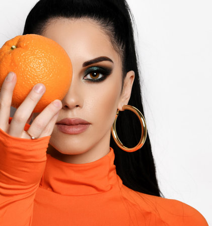 Young brunette woman in modern orange turtleneck sweater, shirt and earrings holds an orange in front of her faceの写真素材