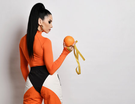 Young woman in bright orange sportswear and turtleneck stands back looking at fresh orange and measuring tape she holdsの写真素材