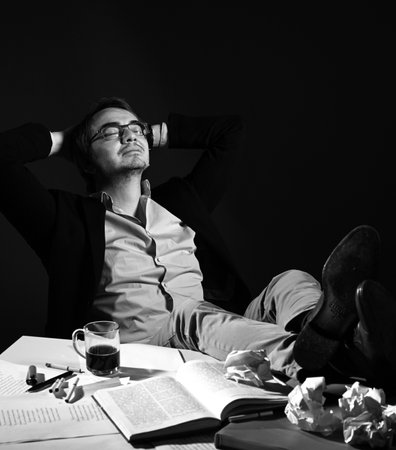 Adult man in casual clothing and glasses sits in dreamy pose with hands behind head, feet on table...