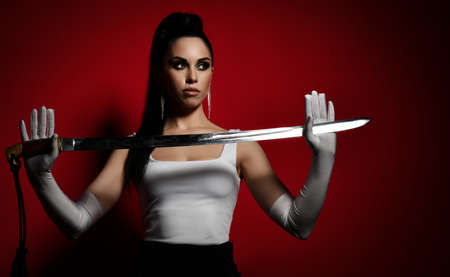 Young pretty brunette woman in white top and gloves standing and holding dagger in hands looking...