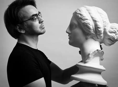 Black and white portrait of adult man in black t-shirt holding in hands, looking at antique sculpture woman headの写真素材