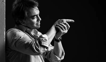 Black and white portrait of adult man in shirt standing back to white concrete wall holding finger gun with hand, aimingの写真素材