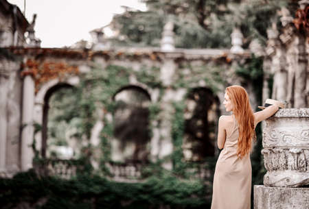 Sensual redhead woman in summer dress stands back to camera at stone flowerpot column in ancient palace gardenの写真素材