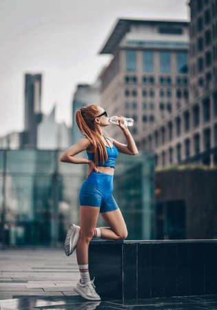 Sporty red-haired woman runner stands at stone bench drinks water during workout on downtown city streetの写真素材