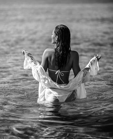 Sexy woman girl in white bikini and shirt goes deeper into the clear sea water. Black and white. Summer vibesの写真素材