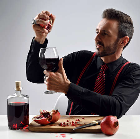Adult man in black shirt with striped tie and suspenders sits at table squeezing fresh pomegranate juice into a glassの写真素材