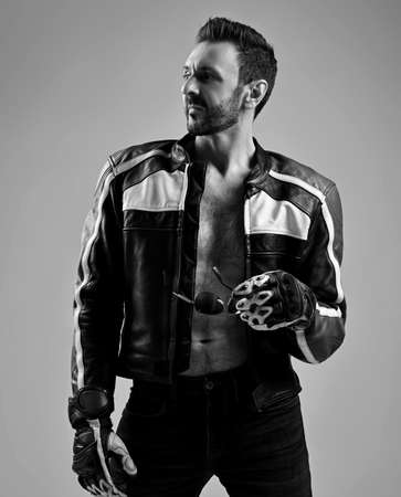 Black and white. Self-confident mature unshaved man in biker leather jacket on naked body stands looking asideの写真素材