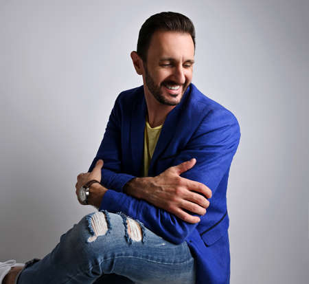 Portrait of laughing stylish adult man in modern torn jeans and blue jacket sitting on stool sidewaysの写真素材