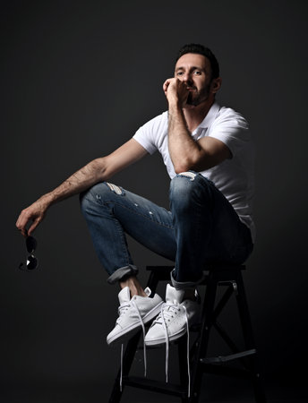 Thoughtful unshaved man in white polo shirt, modern torn jeans and sneakers sits on high chair,...