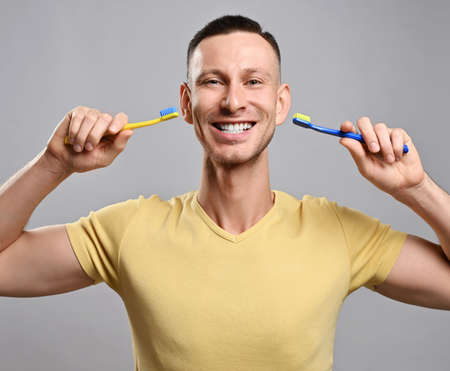 Wide smiling adult man in yellow t-shirt with two toothbrush demonstrates his healthy white teeth....