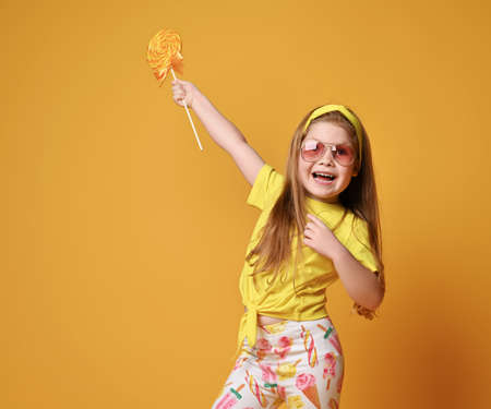 Laughing redhead kid girl in yellow t-shirt, headband, leggings with lollipop pattern stands holding...