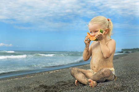 After playing on the sea black sand beach little cute girl picnics of natural vegetarian food - eating a orange slice of fresh ripe papaya, healthy children lifestyle in vacation on the tropical islandの写真素材