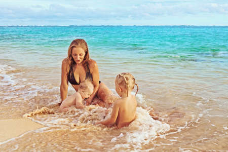 Beautiful smiling young mother with her two little cute children - boy and girl playing joyfully in sea surf on a sand beach, joy of maternity and a healthy happy family on vacation on tropical islandの写真素材
