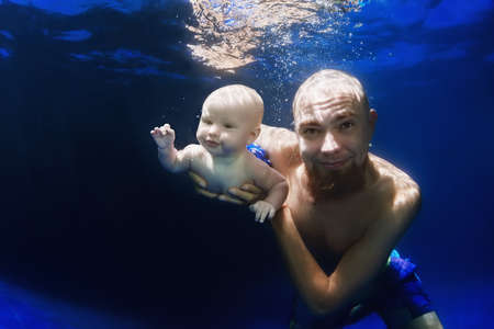 In blue pool father swim underwater with 5 month old baby son - dive deep into clear water with cheerful boy, healthy family lifestyle, child swimming activity and physical growth and developmentの写真素材