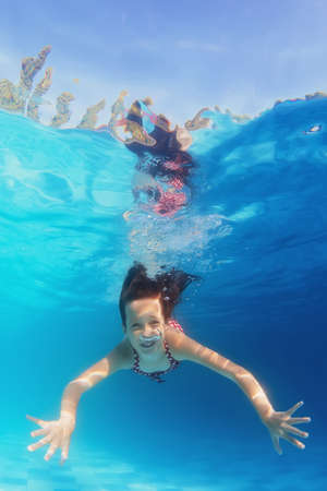 Underwater portrait of happy girl with smiling face swimming in blue pool. Healthy family lifestyle, physical development and children water sports activity with parents on summer vacation with child.の写真素材