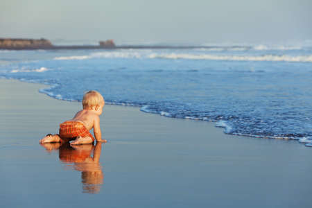On the sunset beach funny baby crawling on black wet sand to sea surf for swimming in the waves. Family lifestyle and water activity during summer vacation with child on the tropical Bali islandの写真素材