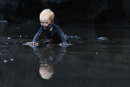 On flat ocean beach dirty baby crawling with fun on wet black sand to sea surf for swimming in waves. Family lifestyle and water activity during summer vacation with child on tropical Bali islandの写真素材