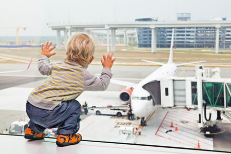 Little baby boy waiting boarding to flight in airport transit hall and looking through the window at airplane near departure gate. Active family lifestyle, travel by air with child on summer vacationのeditorial素材