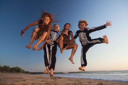 Group young girls in scary skeleton and wild savage costumes jumping high in air with fun before Halloween night party on sunset sea beach. Active people, lifestyles and event celebrations on holidaysの写真素材
