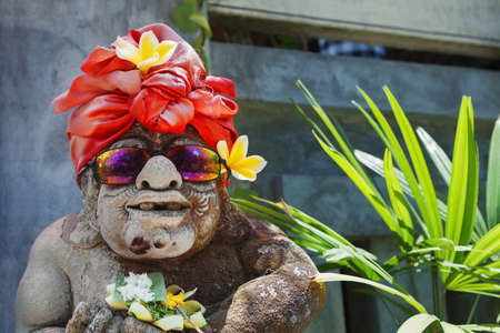 Humorous face portrait of old traditional Balinese temple guard statue dressed in funny tourist costume with sunglasses Arts, culture of Bali and Indonesian people and asian vacation travel backgroundの写真素材