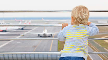 Little baby boy waiting boarding to flight in airport transit hall and looking through the window at airplane near departure gate. Active lifestyle, travel by air with child on family summer vacation.の写真素材