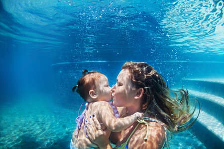 Happy family - mother kiss baby girl dive down with fun in beach pool. Healthy lifestyle, active parents, people water sport activity and underwater swimming lessons on summer vacation with child.の写真素材