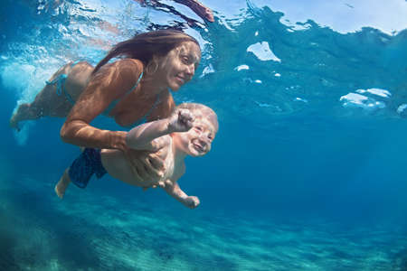 Happy family - mother hold in hands baby girl and dive down with fun in tropical beach sea pool. Active parent lifestyle, people water sport activity, underwater swimming lesson on vacation with childの写真素材