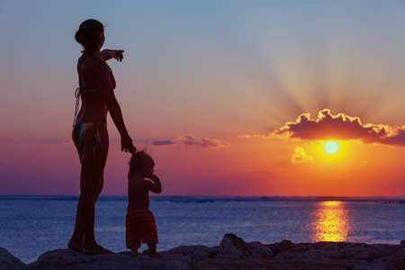 Black silhouette of happy family walking on sea beach - mother hold baby son hand, point sun and look at sunset sky. Active parent lifestyle, outdoor activity on tropical summer vacation with childrenの写真素材