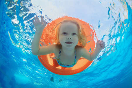 Funny underwater photo of baby girl swimming with fun on orange tube and diving in clear aqua park pool. Healthy family lifestyle, and children water sports activity,  with parents on summer vacation.の写真素材