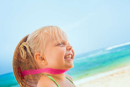 On sunny white sand beach happy smiling baby girl with vietnamese straw hat on head has fun before swimming in sea waves. Active travel family lifestyle, water activity on summer vacation with child.の写真素材
