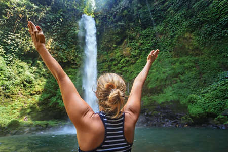 Beautiful young happy girl traveling in Bali wild forest spreading hands enjoying nature under tropical fresh waterfall. Nature day tour, hiking activity adventure and fun on family summer vacation.の写真素材