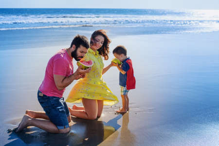 Happy family funny picnic on beach with sea surf - mother feed baby boy, adult man. Father, son eat fruits with fun. Active parents and people outdoor activity on summer vacation with child on Bali.の写真素材