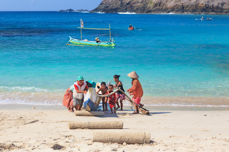 MAWUM BAY FISHER VILLAGE, LOMBOK ISLAND, INDONESIA - OCTOBER 17, 2014 Local children help parents to drag out traditional fisherman boat from turquoise ocean water surf to white sand beach.のeditorial素材
