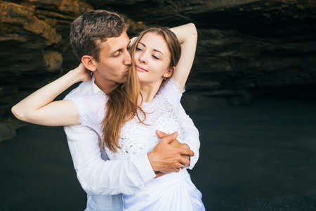 Happy family on honeymoon holiday - just married young man and woman hug with smile on black sand beach with rock background. Active lifestyle, people activity on summer vacation on tropic island.の写真素材