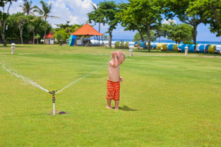 Happy baby boy play with sprinkler spray, swim and splash with fun under water drops on green grass lawn. Travel, active lifestyle, parenting, people activity on summer beach holiday with children.の写真素材
