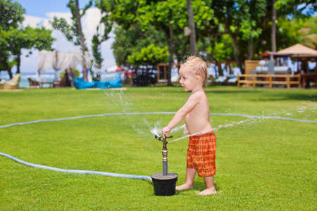 Happy baby boy play with sprinkler spray, swim and splash with fun under water drops on green grass lawn. Travel, active lifestyle, parenting, people activity on summer beach holiday with children.の写真素材