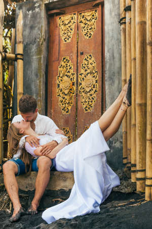 Happy family enjoying romantic honeymoon holiday - just married young man hug and kiss sexy woman lying on black sand beach. Active lifestyle, people activity on summer vacation on tropic island.の写真素材