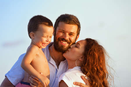 Face portrait of hugging beautiful family - happy mother, father holding baby son on hands walk with fun on sunset beach. Active parents, people outdoor activity on tropical summer holiday with childの写真素材