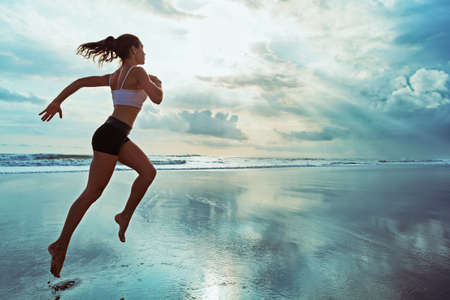 Active sporty woman run along ocean surf by water pool to keep fit and health. Sunset black sand beach background with sun. Woman fitness, jogging workout and sport activity on summer family holiday.