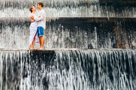 Happy family on honeymoon holidays - married loving couple hugging, kissing with fun under falling water in cascade waterfall pool. Active lifestyle, people travel on summer vacation on Bali island.の写真素材